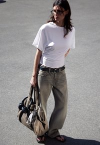 Kvinne som har på seg hvit t-skjorte, løse grå jeans, sort belte og sandaler, mens hun bærer en brun veske med hodetelefoner og tilbehør utendørs.