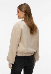 Veste courte beige à texture douce, col mandarin, manches longues bouffantes et taille élastique, associée à un jean noir ajusté.