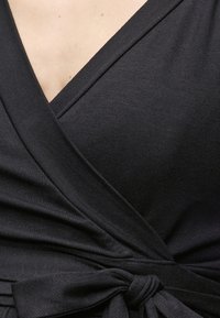 Κοντινή λήψη υφάσματος μαύρου φόρεματος τύπου wrap με διαγώνιες ραφές και δέσιμο φιόγκου στη μέση, αποκαλύπτοντας μερικό ντεκολτέ και δέρμα.