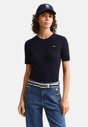 Donna con capelli ricci che indossa una maglia a maniche corte blu navy lavorata a trecce, jeans blu con bottoni dorati, cintura a righe e berretto da baseball blu navy.