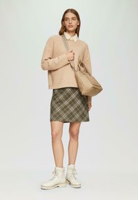 Frau in beigem Pullover, cremigem Hemd, grün kariertem Rock, weißen Schnürboots, die eine sandfarbene Handtasche mit gestreiftem Schultergurt trägt.