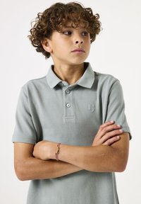 Polo shirt gris clair avec un col, deux boutons et un petit logo sur la poitrine. Le tissu semble doux avec une texture subtile.