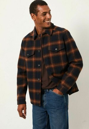 Flanelljacke mit dunkelblauem und orangefarbenem Karomuster, zwei Brusttaschen, Knopfverschluss und gerader Passform, getragen über einem braunen Hemd.