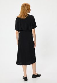 Zwarte midi-jurk met korte mouwen, elastische tailleband en gladde textuur. Draagt zwarte platte schoenen, gezien vanuit de rug tegen een witte achtergrond.