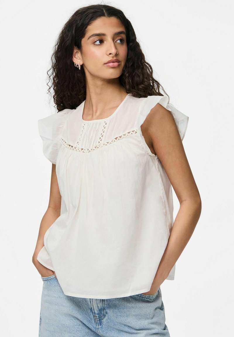 Blusa bianca in cotone con maniche a volants, dettagli in pizzo intricati sulla scollatura, e tessuto leggermente arricciato, abbinata a jeans in denim azzurro chiaro.
