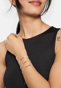 Drei mehrfarbige Perlenarmbänder mit metallischen Akzenten, die an einem Handgelenk getragen werden, vor einem dunklen ärmellosen Oberteil mit glatter Textur.