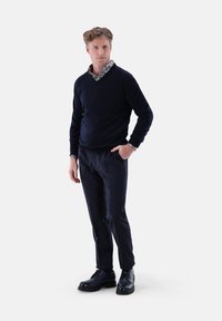 Marineblauer Pullover mit V-Ausschnitt, über einem gemusterten Hemd getragen. Dunkle Hose und schwarze Lederschuhe vervollständigen das Outfit. Minimalistisches Design.