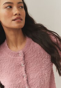 Rožinė, pūkuota kardiganas su apvaliu iškirpimu, papuoštas dekoratyviniais mygtukais ir tekstūruotu paviršiumi. Minkšta medžiaga ir lengvas dizainas.