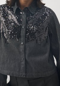 Camicia in denim nera con parte anteriore in paillettes. Presenta un colletto strutturato, chiusura con bottoni e maniche lunghe. Texture liscia con dettagli lucidi.