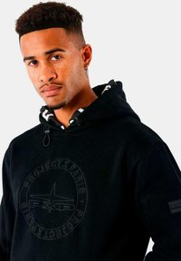 Joven con cabello corto y rizado, vistiendo un hoodie negro con el logo "Project X Paris", anillo en la nariz y arete en forma de cruz, contra un fondo blanco.