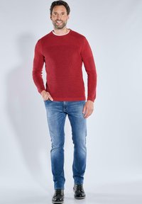 Roter Strickpullover mit gerippten Akzenten, kombiniert mit hellblauen Jeans und schwarzen Schuhen. Das Design betont die Textur und die figurbetonte Silhouette.