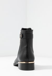 Bottine en cuir noir avec un bout arrondi et un petit talon épais. Dotée d'un accent doré au niveau du talon et d'une bride décorative avec un bouton.