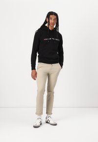 Zwarte hoodie met de tekst "DENIM PROJECT" in wit en rood, gecombineerd met beige broek en witte sneakers met zwarte strepen.