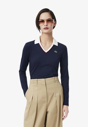 Polo de manga larga en color navy con cuello blanco con ribete rojo y un acento de logo verde. El modelo lleva pantalones de cintura alta en color beige y gafas de sol.
