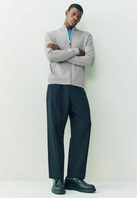 Gray, zip-up sweater with ribbed hem, paired with loose-fitting black trousers and black shoes. Model stands with arms crossed.