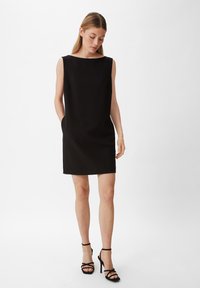 Schwarzes ärmelloses Kleid mit weitem Ausschnitt, zwei seitlichen Taschen und geradem Schnitt, kombiniert mit schwarzen, römischen High Heels.
