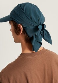 Dunkelblaugrüne Stoffmütze mit einem gebundenen Schal-Detail auf der Rückseite, das eine glatte Textur zeigt. Ein braunes Hemd ist im Hintergrund sichtbar.