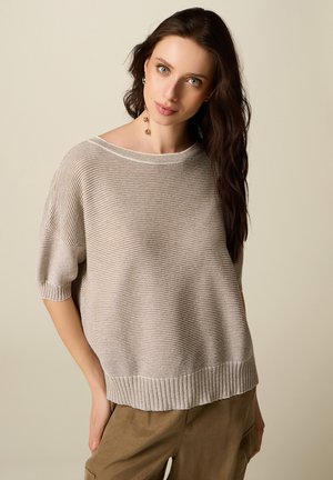 Kvinde med langt mørkt hår iført en løs, beige stribet kortærmet sweater og brune bukser mod en ensfarvet baggrund.