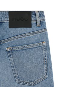 Džinsa bikses ar gaiši zilu mazgāšanas efektu, ar melnu ādas plāksteri, uz kura rakstīts "HUGO", un dubultu šuvi aizmugurējā kabatā.