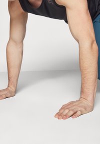 Camisa atlética negra sin mangas en las manos de una persona descansando sobre una superficie blanca, mostrando un tono de piel claro y musculatura definida.