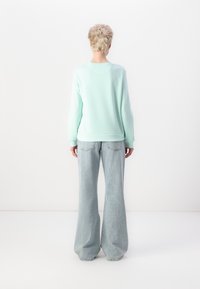 Minta zaļš džemperis ar ribotām piedurknēm un apakšmalu, saskaņots ar gaiši zilām, plašām denim biksēm, kas aprīkotas ar aizmugurējām kabatām. Redzams aizmugurējais skats.