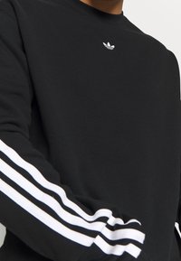 Czarna bluza z długim rękawem wykonana z bawełny, z okrągłym dekoltem, białym wzorem z trzema paskami na rękawach oraz małym białym logo na piersi.