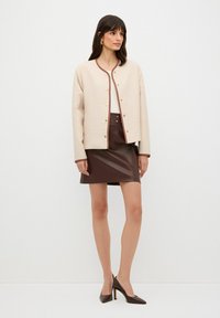 Beige vadderad jacka med guldknappar och brunt kant, tillsammans med en brun läderminikjol och bruna spetsiga klackar. Släta texturer, modern passform.