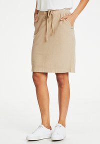 Jupe beige jusqu'aux genoux, en tissu doux et texturé, dotée d'une taille élastique avec cordon de serrage et de poches latérales. Associée à des baskets blanches.