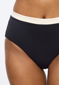 Κοντινό πλάνο ατόμου που φοράει μαύρο μαγιό με ψηλή μέση και λευκή μέση, εστιάζοντας στη μέση και την άνω περιοχή του μηρού.