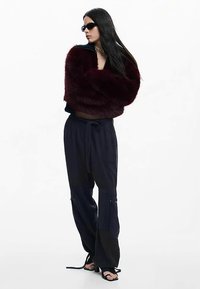 Veste en fausse fourrure bordeaux, design court, associée à un pantalon cargo noir avec des fermetures éclair. Portée avec des lunettes de soleil noires et des sandales à brides.