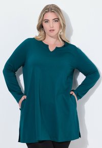 Türkisfarbene Langarm-Tunikabluse mit V-Ausschnitt, seitlichen Schlitzen und Taschen. Hergestellt aus einem glatten, dehnbaren Stoff für hohen Komfort.