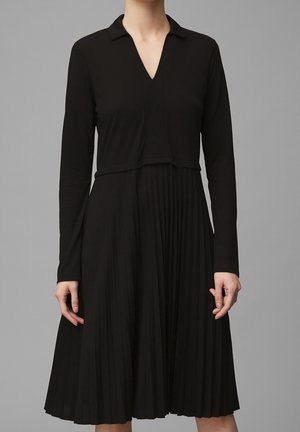 Schwarzes langärmeliges Kleid mit Kragen, V-Ausschnitt und plissiertem Rock. Der Stoff wirkt glatt und fließend.