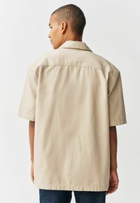 Chemise beige à manches courtes en tissu léger, dotée d'un col plat et d'une coupe décontractée avec une texture lisse. Vue arrière montrée.