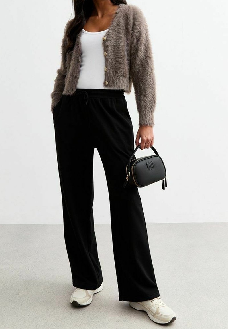 Femme portant un cardigan gris duveteux sur un haut blanc, un pantalon large noir, des baskets blanches, tenant un petit sac à main noir.