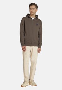 Joven de pie, vestido con una sudadera marrón con cremallera, pantalones color crema y zapatillas beige contra un fondo claro y liso.