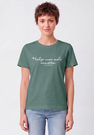 Junge Frau mit kurzen braunen Haaren, die ein grünes T-Shirt mit weißem deutschem Text und hellblaue Jeans trägt, steht vor einem schlichten weißen Hintergrund.