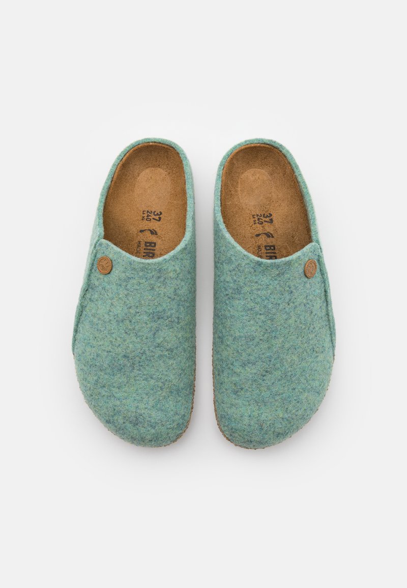 Sabots en feutre de couleur turquoise clair avec une texture lisse et un accent en forme de bouton rond. Comprend des semelles en liège marron et des semelles en caoutchouc.