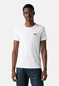Ung mand står oprejst iført en hvid crew neck Levi's t-shirt med rødt logo på brystet og mørkeblå jeans foran en ensfarvet baggrund.