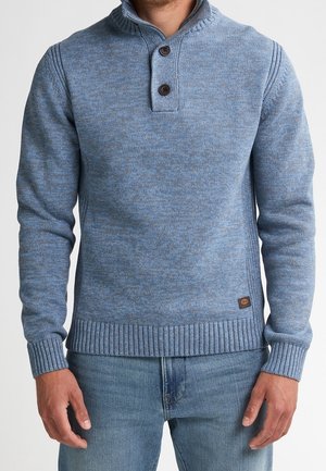 Mann trägt einen hellblauen Strickpullover mit hohem Rippenkragen und zwei braunen Knöpfen, kombiniert mit blauen Jeans, im Bildausschnitt von Hals bis Hüfte.