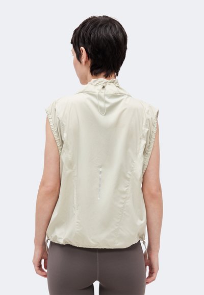 Άτομο με το πρόσωπο προς τα πίσω, που φοράει αμάνικο μπεζ μπουφάν με σούρα στο γιακά και κορδόνι στο τελείωμα πάνω από σκουρόχρωμες κολάν.