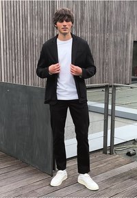 Ung man som justerar en svart blazer över en vit t-shirt, bärande svarta byxor och vita sneakers, stående på en trädäck vid en modern byggnad.