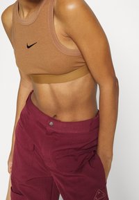 Brun ribbad sportbh med en svart Nike-logga, kombinerad med maroon cargobyxor med sidofickor och knappstängning.