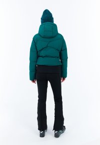 Žmogus, vilkintis mėlyną pūkų striukę, mėlyną megztinę, juodas kelnes ir juodus slidinėjimo batus, stovi atsisukęs atgal priešais baltą foną.