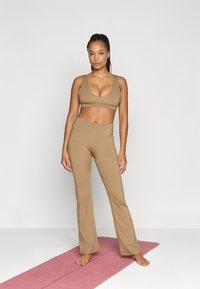 Ταν αθλητικό crop top και φαρδιές παντελόνες από λείο ύφασμα, φορεμένα με γυμνά πόδια σε ροζ στρώμα γιόγκα σε απλό φόντο.