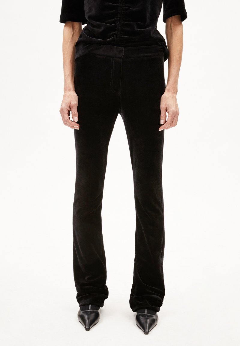 Pantaloni in velluto nero con silhouette aderente, leggera elasticità e orlo svasato, abbinati a tacchi alti neri a punta. Texture liscia.