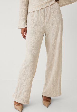 Kvinde iført beige teksturerede bukser med vide ben og matchende langærmet top, kombineret med beige spidse hæle med små spænde-detaljer.