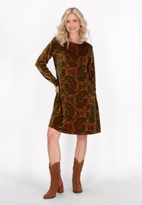 Zöld és narancssárga paisley mintás bársonydressz, hosszú ujjú, laza fazonú, térd hosszúságú, barna velúr bokacsizmával párosítva.