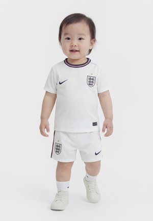 Tout-petit portant un kit de football blanc de l'Angleterre avec des logos Nike, debout et souriant sur un fond uni.