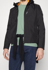 Svart lättviktsjacka med huva och ett orangefärgat innerfoder, med fickor som stängs med dragkedja och en stilren, modern design. Bärs över en grön skjorta.