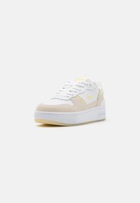 Hvit sneaker med beige semsket skinndetaljer, flate snørebånd og en teksturert gummisåle. Har en diskret gul detalj og merkevarelogo på siden.